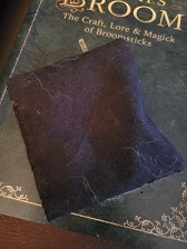 A small purple sachet bag, laying atop a book entitled The Witch’s Broom: The Craft, Lore & Magick of Brooms