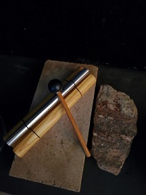 a silver cylinder with a wooden mallet resting upon a stone rectangle, with a fist-sized rock on the right side 