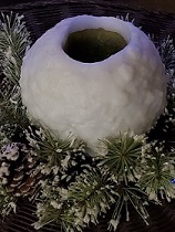 a round white candle shaped like a snowball surrounded by pine boughs